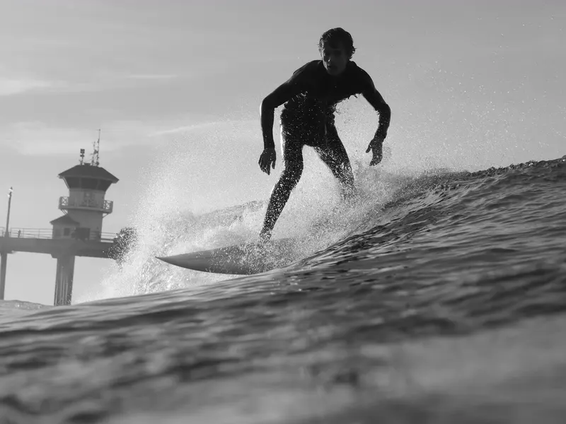 A surfer in a wetsuit riding a small wave, with the Huntington Beach Pier in the background